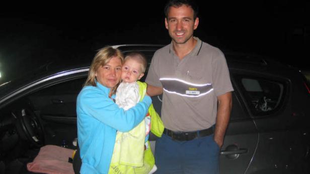 Eine Frau mit einem Baby im Arm und ein Mann stehen vor einem Auto.