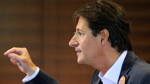 Ein Mann mit Brille gestikuliert vor einem braunen Hintergrund.