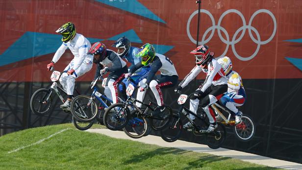 Mehrere BMX-Fahrer starten ein Rennen vor dem Hintergrund der Olympischen Ringe.