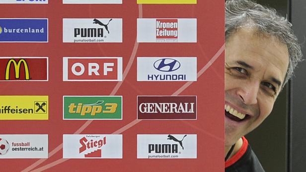 Ein Mann lächelt hinter einer Wand mit Sponsorenlogos hervor.