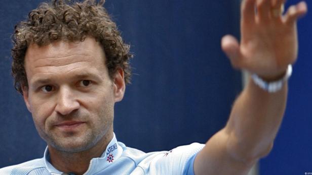 Ein Mann mit lockigem Haar hebt die Hand zum Gruß.