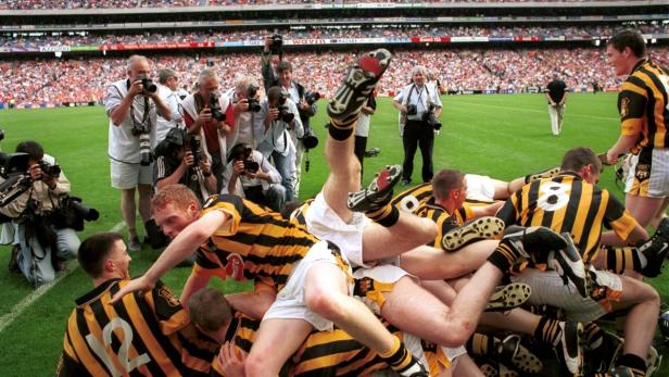 Hurling-Spieler feiern einen Sieg auf dem Spielfeld, umringt von Fotografen.