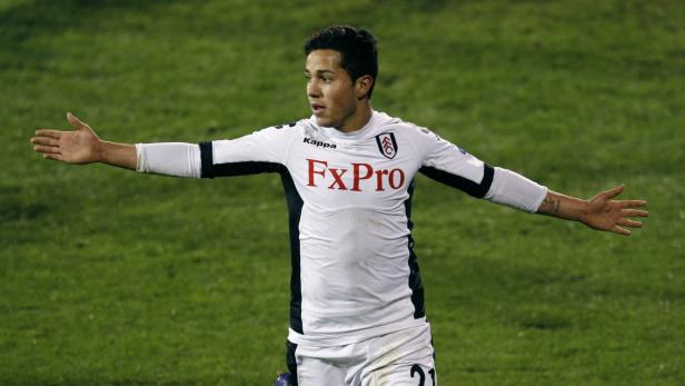 Ein Fußballspieler im weißen Trikot mit der Aufschrift „FxPro“ steht mit ausgestreckten Armen auf dem Spielfeld.