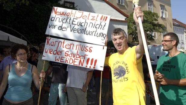 Menschen demonstrieren mit Schildern, auf denen „Neuwahl Jetzt“ und „Kärnten muss wieder sauber werden“ steht.