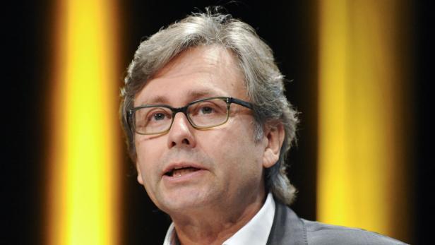 Ein Mann mit Brille und grauem Haar spricht vor einem gelben Hintergrund.