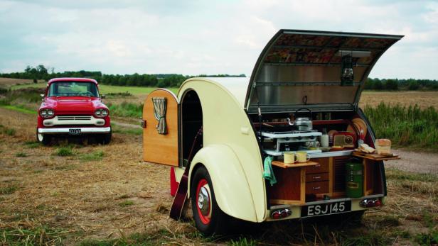 Ein roter Oldtimer-Truck und ein Wohnwagen mit offener Heckklappe auf einem Feld.