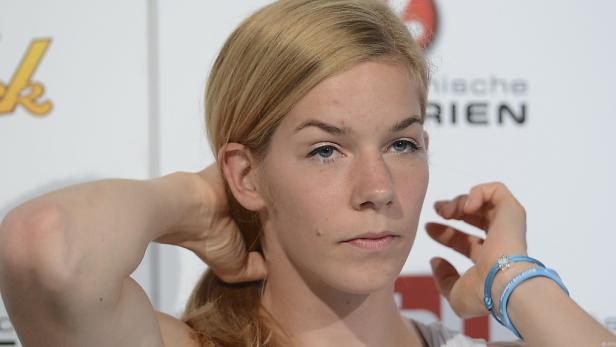 Eine blonde Frau mit blauen Armbändern posiert vor einer Wand.