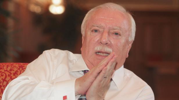 Ein Mann mit weißem Haar und Schnurrbart hält die Hände vor dem Gesicht.