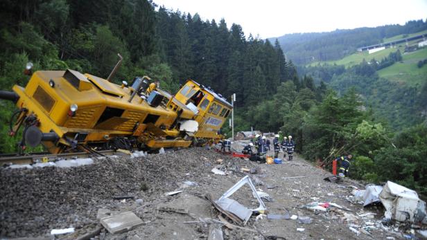 Ein entgleister, gelber Zug liegt auf der Seite, während Rettungskräfte in der Nähe stehen.
