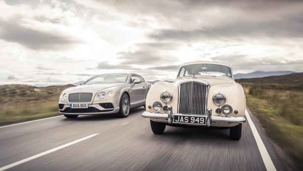 Zwei Bentley-Fahrzeuge, ein Oldtimer und ein modernes Modell, fahren auf einer Landstraße.