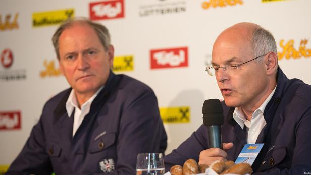 Zwei Männer in Trachtenjacken sitzen vor einer Wand mit Tirol-Logos.