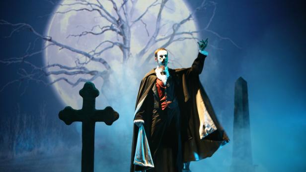 Ein als Dracula verkleideter Mann steht auf einer Bühne vor einem Vollmond.