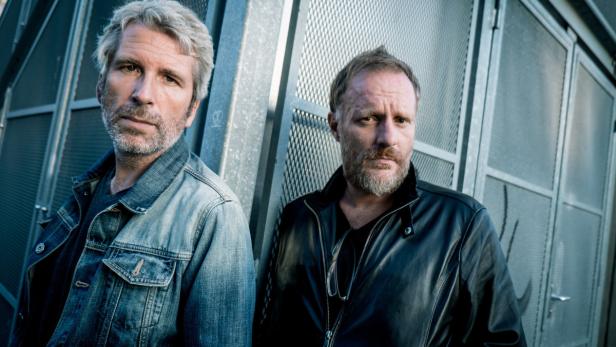 Zwei Männer posieren vor einer Wand mit Metallgitter.