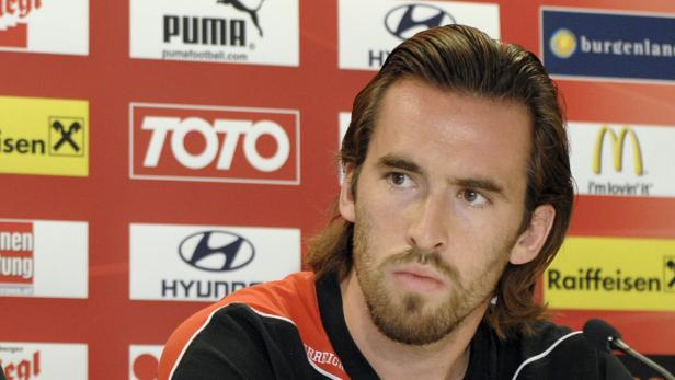 Ein Mann mit Bart vor einer Wand mit Sponsorenlogos wie Puma, Toto und McDonald's.