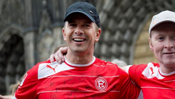 Ein lächelnder Mann mit Kappe und einem signierten Trikot von Fortuna Düsseldorf.