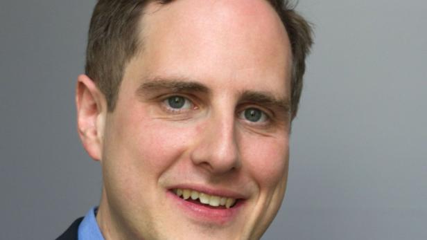 Ein lächelnder Mann mit hellbraunen Haaren und blauen Augen vor einem grauen Hintergrund.