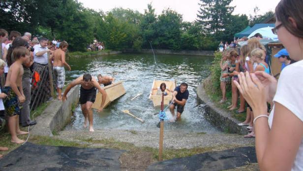 Zuschauer beobachten ein Rennen mit selbstgebauten Booten auf einem kleinen See.