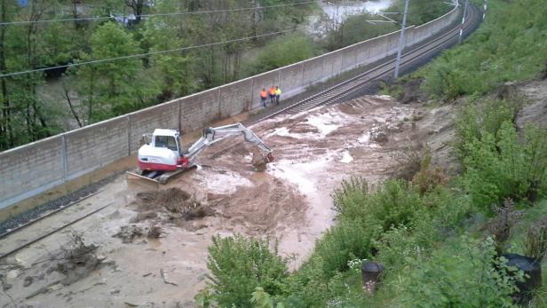 Ein Bagger räumt Schlamm von einer Bahnstrecke, während Arbeiter in Warnwesten zusehen.