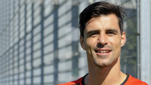 Ein lächelnder Mann mit dunklen Haaren vor einer modernen Fassade.