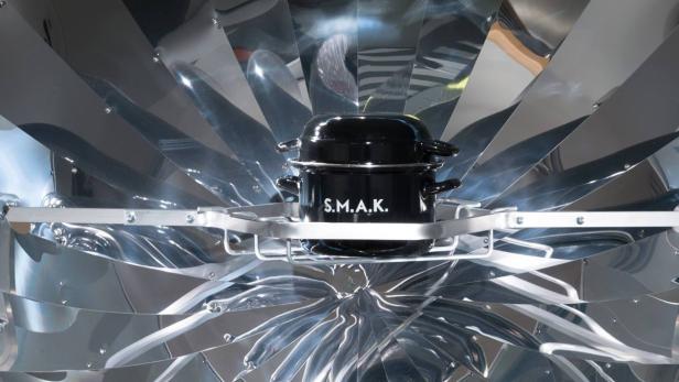 Ein schwarzer Kochtopf mit der Aufschrift S.M.A.K. steht in einem Solarkocher.