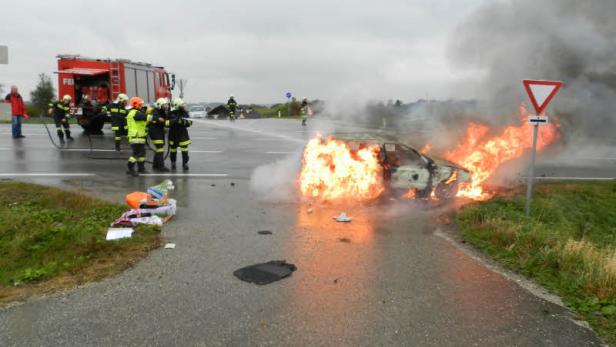 Ein brennendes Auto steht auf einer Kreuzung, während Feuerwehrleute den Brand bekämpfen.