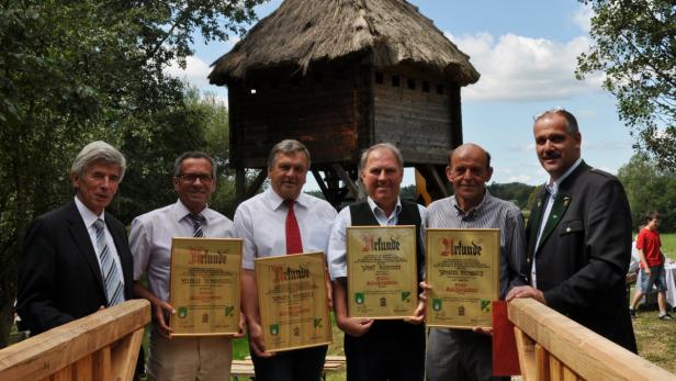 Eine Gruppe von Männern präsentiert Urkunden vor einem hölzernen Turm mit Strohdach.