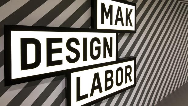 Leuchtschilder mit der Aufschrift „MAK Design Labor“ an einer gestreiften Wand.