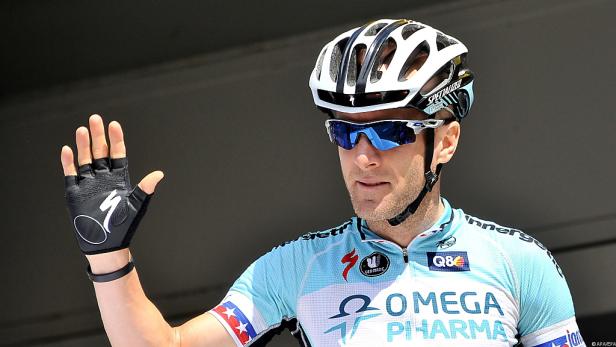 Ein Radrennfahrer mit Helm und Sonnenbrille winkt in die Kamera.