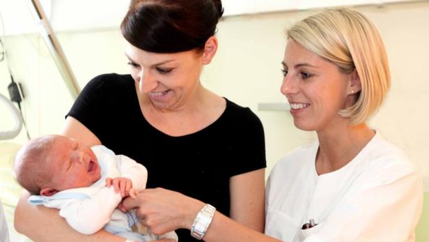 Eine Frau hält ein schreiendes Baby, während eine Krankenschwester lächelt.