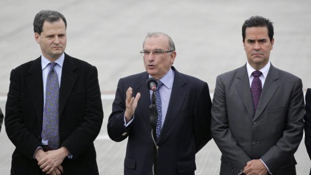 Drei Männer in Anzügen stehen nebeneinander, einer spricht in ein Mikrofon.