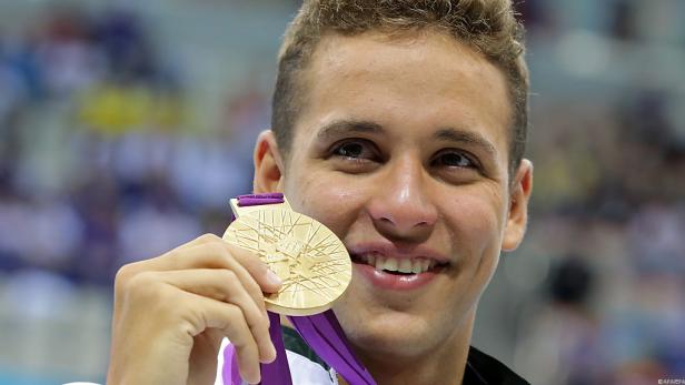 Ein junger Mann lächelt und hält stolz seine Goldmedaille in der Hand.