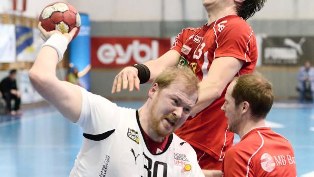 Ein Handballspieler in Weiß holt zum Wurf aus, während zwei Gegenspieler in Rot ihn bedrängen.