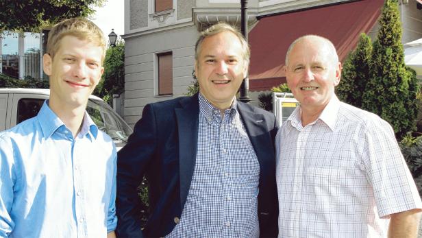 Drei Männer unterschiedlichen Alters stehen lächelnd vor einem Gebäude.