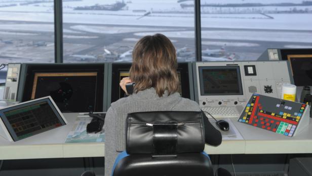 Eine Fluglotsin sitzt vor mehreren Bildschirmen und einem Fenster mit Blick auf einen Flughafen.