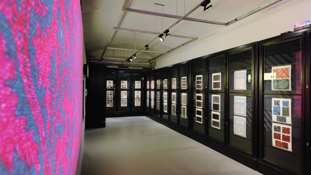 Ein Ausstellungsraum mit Mustern in Vitrinen und einer farbenfrohen Wand im Vordergrund.