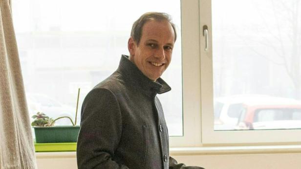 Ein lächelnder Mann in einem grauen Mantel steht vor einem Fenster.