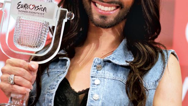 Conchita Wurst hält eine Eurovision-Trophäe und lächelt.