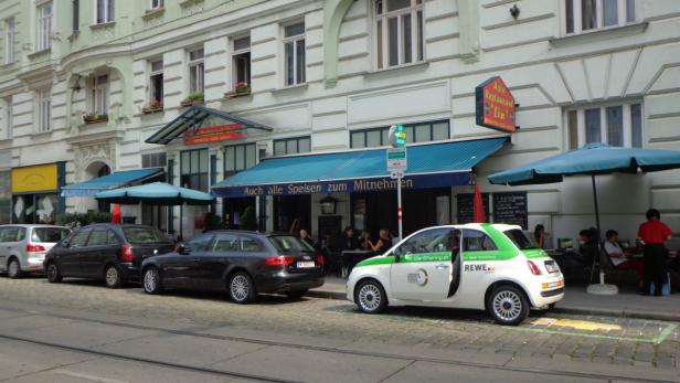Ein belebtes Straßenbild mit geparkten Autos und einem Restaurant mit Außengastronomie.