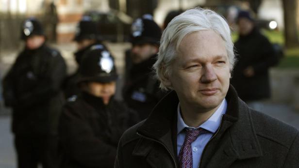 Julian Assange mit hellgrauem Haar und dunklem Mantel, umgeben von Polizisten.