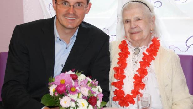 Ein Mann im Anzug mit einem Blumenstrauß sitzt neben einer älteren Frau mit Blumenkette.