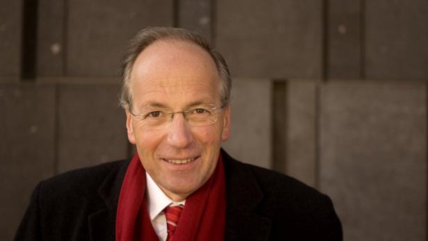 Ein lächelnder Mann mit Brille, Schal und dunklem Mantel vor einer Wand.