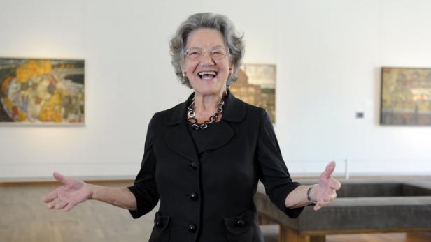 Eine ältere Frau mit Brille und schwarzem Blazer steht lächelnd in einer Kunstgalerie.