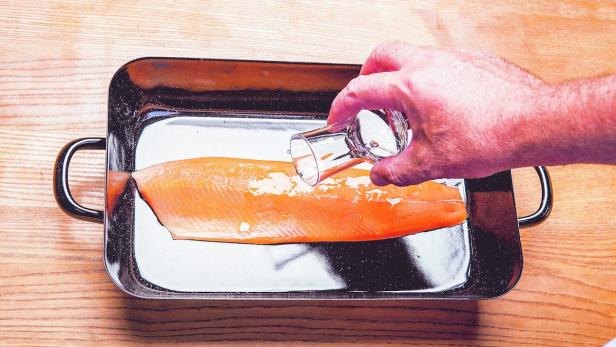 Eine Person beträufelt ein Lachsfilet in einer schwarzen Pfanne mit einer klaren Flüssigkeit.