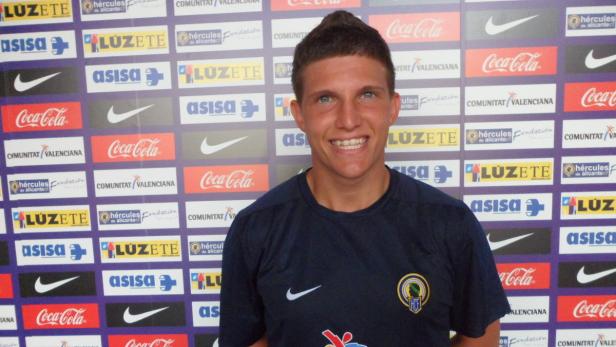 Ein junger Mann lächelt vor einer Wand mit Logos von Sponsoren wie Coca-Cola und Luzete.