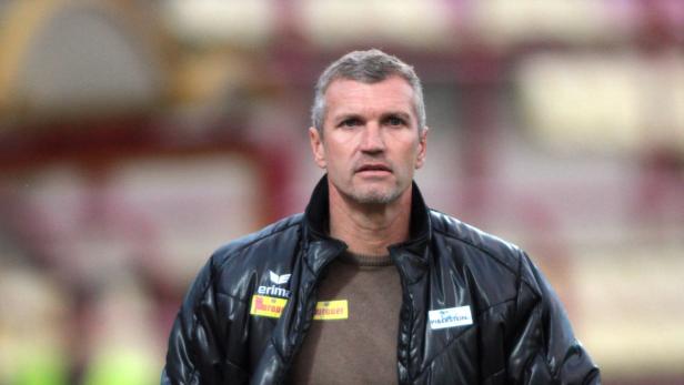 Ein Mann mit grauen Haaren und einer schwarzen Jacke steht vor einem unscharfen Hintergrund.