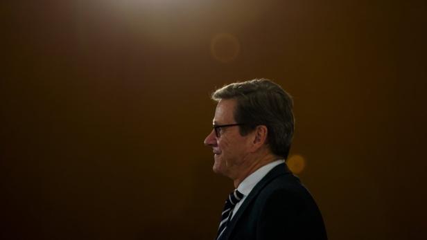 Ein Mann mit Brille und Anzug im Profil vor einem dunklen Hintergrund.