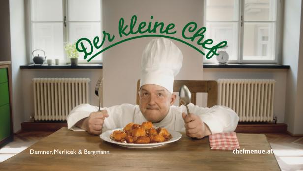 Ein Koch mit Kochmütze präsentiert ein Gericht mit Gabel und Löffel.