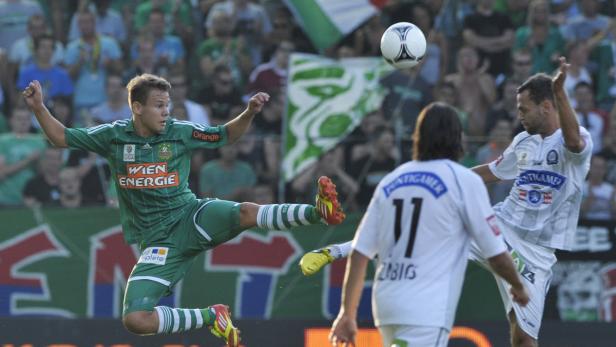 Ein Fußballspieler in Grün springt, um den Ball zu treten, während ein anderer Spieler in Weiß zuschaut.