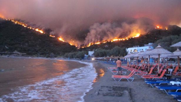 Ein Waldbrand wütet auf einem Hügel hinter einem Strand mit Liegestühlen.