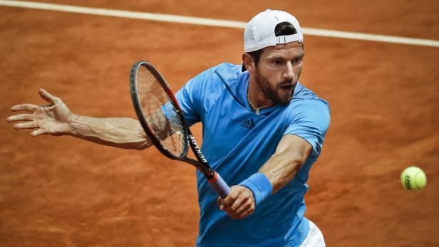 Ein Tennisspieler in blauem Hemd und weißer Kappe schlägt einen Ball.
